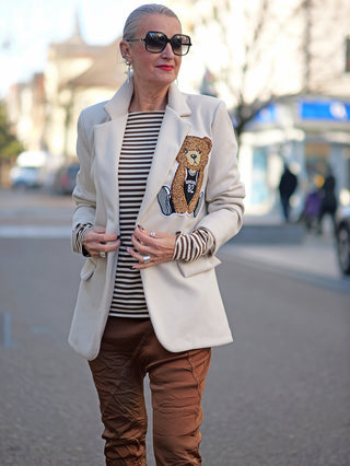 Frau auf der Straße trägt OFF#DLY Blazer Bear creme mit großem Bären-Patch, Sonnenbrille und braune Hose. Ihre Hände sind sichtbar, während sie im modischen Streetwear-Stil geht.
