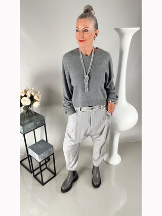 Frau trägt OFF#DLY Stripes Pant Lola grey, eine lässige Bundfaltenhose mit tiefem Schritt und seitlichen Taschen, kombiniert mit grauem Pullover und grauen Stiefeln, neben einer Vase mit weißen Rosen.