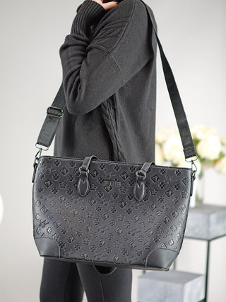 Person hält den Sweet Years Shopper schwarz, einen großen schwarzen Shopper mit geprägtem Design, Reißverschluss, Henkeln und verstellbarem Riemen. Zwei Innenfächer und ein Rückseitenfach mit Reißverschluss sind sichtbar.
