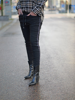 OFF#DLY Pant Tina black: Person trägt schwarze Joggpants mit seitlicher Kettenkordel und schwarzer Flanellbluse, kombiniert mit schwarzen Stiefeln. Ideal für lässige Straßenmode und bequemen Alltagsstil.