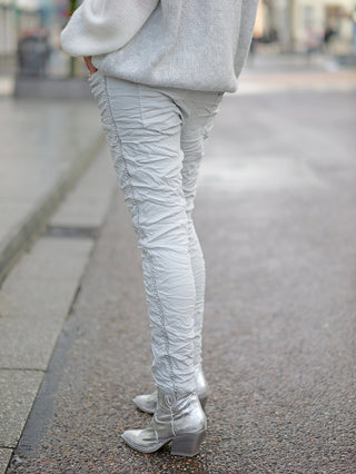 Eine Person trägt die OFF#DLY Pant Tina grey, eine leichte Joggpant mit Kettenkordel und Gummibund, kombiniert mit silbernen Schuhen, sichtbar auf einem Steinweg.