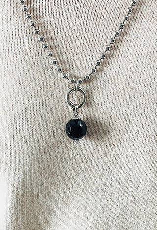 Nahaufnahme der Glam & Rock Onyx Kugel: ein silbernes Kugelkettchen mit einem polierten Silberring, von dem ein einzelner facettierter schwarzer Onyx-Anhänger an einem beigen, gerippten Strickstoff hängt.