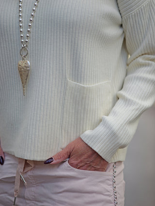Person trägt den OFF#DLY Rippulli Renate beige, ein schmaler Ripppulli mit aufgesetzter Tasche, begleitet von einer Halskette.