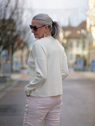 Frau in weißem OFF#DLY Rippulli Renate Pullover mit Tasche, trägt Sonnenbrille und rosa Hose. Pullover hat eine schmale, gerade Passform.