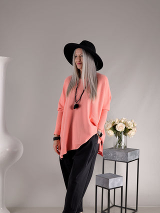 Person trägt den OFF#DLY Pulli Olivia apricot, einen locker sitzenden, korallfarbenen Strickpullover mit geradem Schnitt, kombiniert mit schwarzem Hut, schwarzen Hosen und schwarzem, geschichtetem Schmuck.