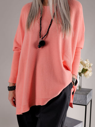 Person in locker sitzendem OFF#DLY Pulli Olivia apricot, kombiniert mit schwarzen Hosen. Der Pullover hat lange Ärmel und wird mit schwarzen Accessoires ergänzt, einschließlich einer langen Halskette und Armbändern.