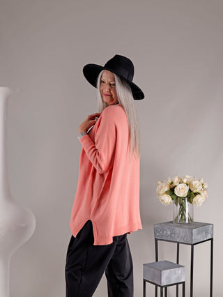 Eine reife Frau trägt den OFF#DLY Pulli Olivia apricot in Korallenrosa mit schwarzen Hosen und einem breiten Hut, ergänzt durch silbernes Haar und eine Vase mit weißen Rosen im Hintergrund.