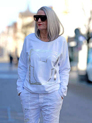 Frau trägt das OFF#DLY Longsleeve Frame weiß mit Silberprint und Rundhalsausschnitt, kombiniert mit Sonnenbrille, in einer urbanen Umgebung.