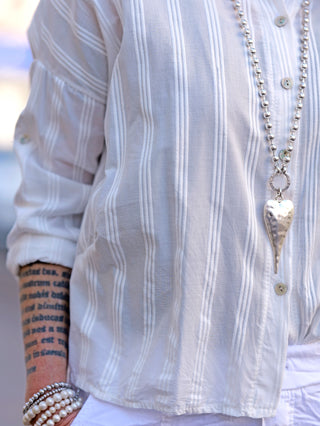OFF#DLY Bluse Vera weiß getragen von Person; Streifenbluse mit Rundhalsausschnitt und Knopfleiste, hinten länger geschnitten. Sichtbare Details: Arm mit Schriftzug und Kette auf dem Shirt.