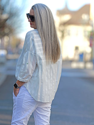Die OFF#DLY Bluse Vera weiß, getragen von einer Frau mit langen grauen Haaren und Sonnenbrille, zeigt einen Rundhalsausschnitt und Knopfleiste, ideal für lässige Street-Fashion.