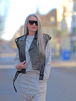 Eine Frau trägt die OFF#DLY Fake Leder Biker Weste mit seitlichen Taschen und Gürtel, kombiniert mit Sonnenbrille, in einem modischen Streetwear-Look.