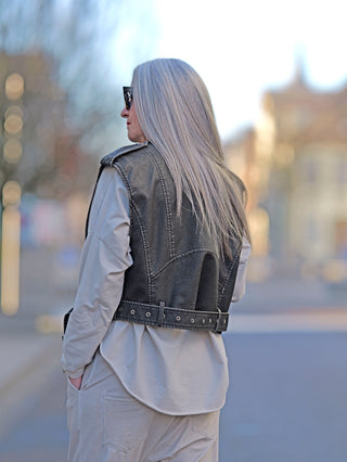 Frau mit langer Haarpracht trägt die OFF#DLY Fake Leder Biker Weste mit seitlichen Taschen und Gürtel. Die Weste hat einen geraden Schnitt und ist in Größe S/M abgebildet.