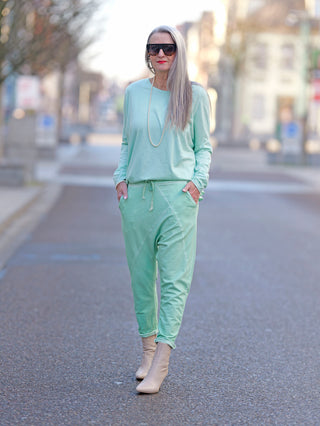Frau trägt OFF#DLY Jogger Pant Tina türkis, Baggy-Stil, mit Gummibund und Kordelzug, seitlichen Taschen. Kombiniert mit Sonnenbrille und Perlenkette, betont lässigen Street-Fashion-Look.