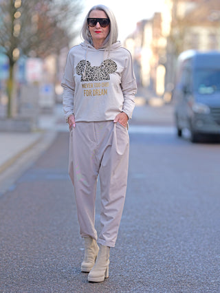 Frau trägt den OFF#DLY Hoody Tee Dream beige mit Mausdesign und Goldprint, während sie auf der Straße geht. Der Hoodie hat eine gerade Passform und lange Ärmel.