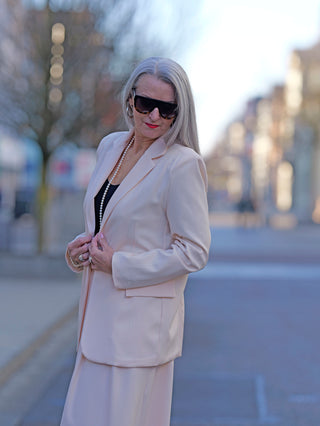 Frau trägt OFF#DLY Blazer Bibi apricot, ein leichter, gefütterter Ein-Knopf-Blazer mit angedeuteten Taschen, Sonnenbrille und Perlenkette, präsentiert urbanen Modestil.