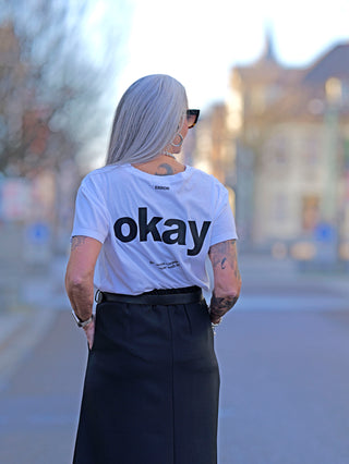 Frau mit Tattoos trägt das Liv Bergen Tee OKAY in optic white, ein gerades Rundhals-T-Shirt mit Rückenprint. Sie kombiniert es mit einem schwarzen Rock, im Outdoor-Setting.