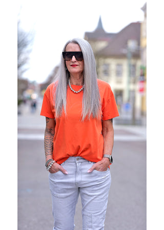 Frau mit grauen Haaren trägt das Liv Bergen Tee OKAY arancia in Orange, mit Rundhalsausschnitt und Rückenprint. Sie trägt es lässig mit Jeans, Accessoires und Sonnenbrille.