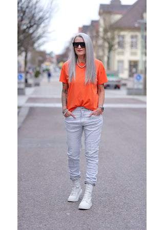 Frau auf der Straße in Liv Bergen Tee OKAY arancia, orangefarbenes T-Shirt mit Rundhals und Rückenprint, kombiniert mit Jeans und weißen Stiefeln. Perfekt für einen lässigen, modischen Alltagslook.