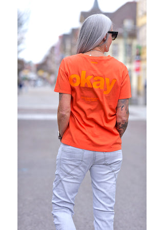 Liv Bergen Tee OKAY arancia, getragen von einer Person mit Tattoos, zeigt einen Rundhalsausschnitt und Rückenprint. Das Model trägt Jeans und eine Sonnenbrille. Perfekt für einen lässigen Look.