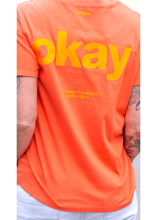 Person trägt das Liv Bergen Tee OKAY arancia, ein oranges T-Shirt mit gelbem Schriftzug. Es hat einen Rundhalsausschnitt und ist aus 100% Baumwolle, mit geradem Schnitt.