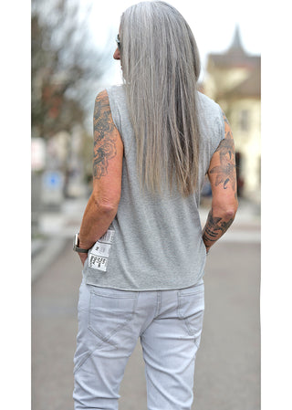 Person trägt Liv Bergen Top EAST Melange mit Rundhalsausschnitt und kleinen Patches, kombiniert mit weißen Hosen. Das Top hat eine gerade Passform und besteht aus Tencel und Baumwolle.