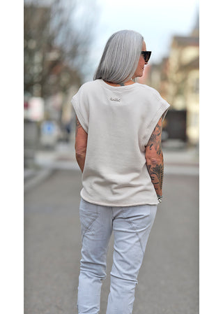 Frau mit Tattoos trägt Liv Bergen V Shirt dirty white, ärmelloser Sweater mit V-Ausschnitt, leicht oversized, 3D Emblem am Rücken, kombiniert mit weißen Hosen.