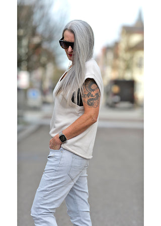 Frau mit langen grauen Haaren trägt Liv Bergen V Shirt dirty white, ärmelloser Sweater mit V-Ausschnitt, sichtbarem 3D-Emblem am Rücken, gerade leicht oversized Passform, kombiniert mit weißer Hose und Sonnenbrille.