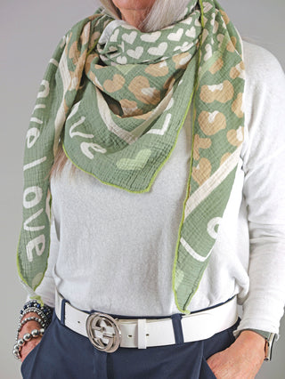 Frau trägt das OFF#DLY Tuch LOVE khaki, ein stilvolles Musselintuch mit Print. Sichtbar sind auch weiße Gürtel mit silberner Schnalle und Armbänder.