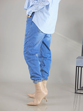 Person trägt OFF#DLY Denim Jogger in Midblue, mit Gummibund, Kordelzug, großen Doppeltaschen vorne und zwei Taschen hinten. Kombiniert mit Boots und einem blau-weiß gestreiften Shirt.