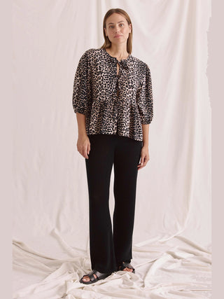 Frau trägt die Cotton Candy Bluse Mina Multicolor mit Leopardenprint, Schnürung vorne und Puffärmeln, kombiniert mit lässigen Hosen und schwarzen Sandalen.