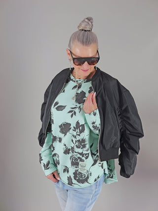 OFF#DLY Longsleeve Flower mint/schwarz gezeigt an einer Frau mit Sonnenbrille, schwarzer Jacke und blumigem Boxy-Sweatshirt mit Rundhalsausschnitt. Perfekt für lässige, stilvolle Looks. Größenempfehlung bis 44.
