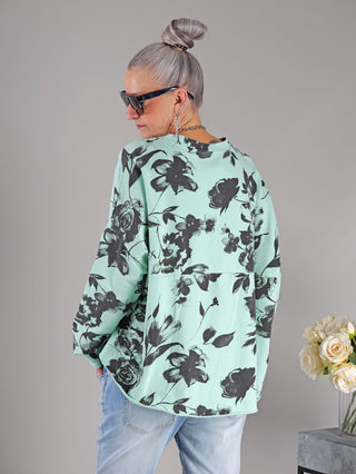Frau trägt OFF#DLY Longsleeve Flower mint/schwarz mit floralem Muster und Rundhalsausschnitt. Boxy Passform sichtbar, kombiniert mit Sonnenbrille. Ideal für Größen bis 44.