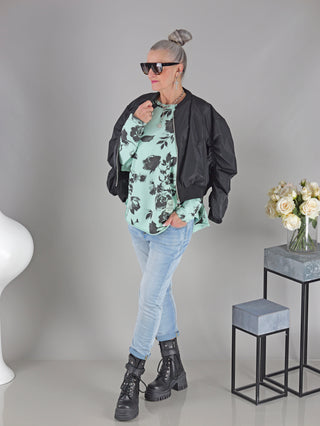 Frau trägt OFF#DLY Longsleeve Flower mint/schwarz, ein Boxy Sweatshirt mit floralem Print und Rundhalsausschnitt, kombiniert mit schwarzer Jacke und Sonnenbrille.