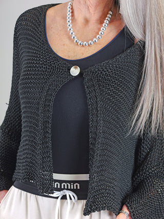 Frau trägt die OFF#DLY Jacke Manuela black, eine Grobstrickjacke mit Einknopfverschluss aus 100% Baumwolle. Sichtbar ist ein silberner Halsketten-Akzent über dem schwarzen Pullover.