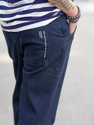 Person trägt Nuomeno Denim Carrot blue Jeans mit seitlichen Einschubtaschen und großem Logo auf den hinteren Taschen. Sichtbar sind eine blaue und weiße gestreifte Hemd und ein Handgelenk-Tattoo.