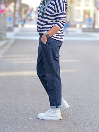 Person trägt Nuomeno Denim Carrot blue Hose mit seitlichen Einschubtaschen, großem Logo auf den Gesäßtaschen, und Gummibund hinten. Kombiniert mit gestreiftem Oberteil und weißen Schuhen.