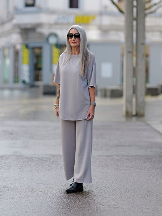 Frau trägt OFF#DLY Loungewear Set taupe, graues Outfit mit 3/4-Ärmeln und seitlichen Taschen, kombiniert mit Sonnenbrille und schwarzen Stiefeln, im Straßenmodus.
