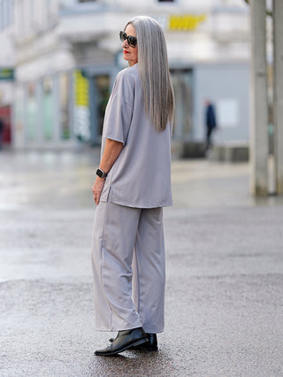 Frau in grauem OFF#DLY Lougewear Set taupe mit 3/4 Ärmeln und seitlichen Taschen, schwarze Stiefel, trägt Sonnenbrille und Uhr, steht auf einer Straße.
