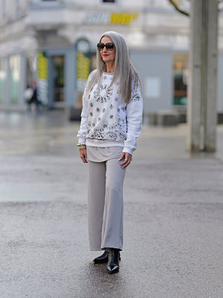 Frau trägt OFF#DLY Sweater Marisol offwhite mit Paillettenstickerei, kombiniert mit Sonnenbrille und grauer Hose, auf der Straße. Nahaufnahme zeigt schwarze Stiefel.
