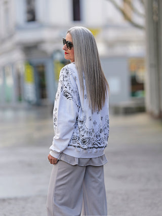 Frau mit grauem Haar trägt den OFF#DLY Sweater Marisol offwhite mit Print und Pailletten-Stickerei, kombiniert mit grauer Hose, in urbanem Umfeld.