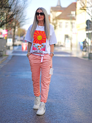 Frau trägt das OFF#DLY Tee Sun white mit Sonnen- und Wolkendesign, kombiniert mit rosa Hose und weißen Sneakers. Kurzarmlänge, gerade Passform, verziert mit Kunststoffperlen.