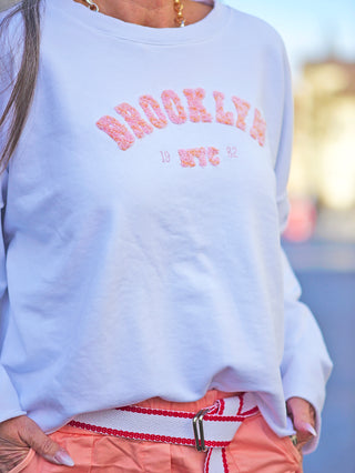 Person trägt „OFF#DLY Sweat Brooklyn white“: weißes Sweatshirt mit rundem Halsausschnitt und Teddy-Print „Brooklyn“. Gerade Passform, aus Baumwolle-Elastan-Mischung. Perfekt für lässige Anlässe.