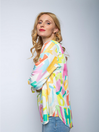 Emily Bluse 8564-152710 multi/letters: Frau mit blonden Haaren trägt eine farbenfrohe Shirtbluse mit Stehkragen und langen Ärmeln. Fließender Viskose-Satin mit Letterprint und Streifenmanschetten.