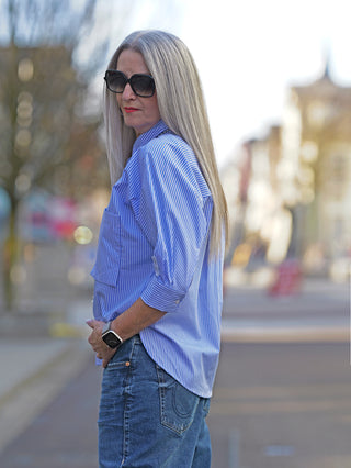 Nuomeno Concept Bluse Corta bianco, getragen von einer Frau mit langen Haaren und Sonnenbrille. Die Bluse hat weite 3/4 Ärmel, Knopfleiste und große Brusttasche.