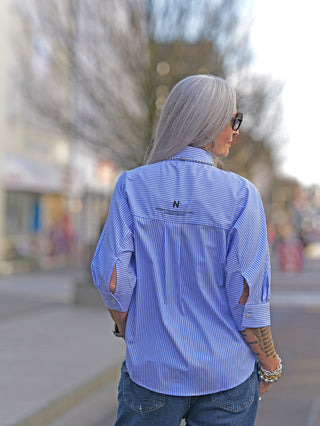 Frau mit grauen Haaren und Tattoos trägt die Nuomeno Concept Bluse Corta bianco, eine stilvolle Boxy-Bluse mit weiten 3/4 Ärmeln, Knopfleiste und großer Brusttasche.