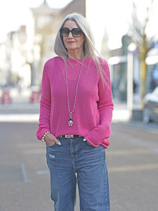 Frau trägt Liv Bergen Crochet Sweater Pink mit langen Ärmeln und weitem Schnitt. Der Netzstoff zeigt den Text LVBRGN auf der Rückseite. Kombiniert mit Jeans und Sonnenbrille.