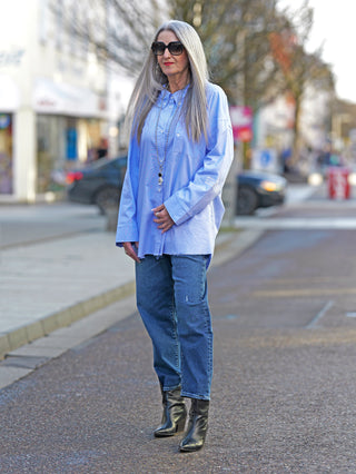 Frau auf dem Bürgersteig trägt die Nuomeno Concept Bluse Azzurro mit Knopfleiste und gedrehter Brusttasche, kombiniert mit Jeans und Stiefeln.