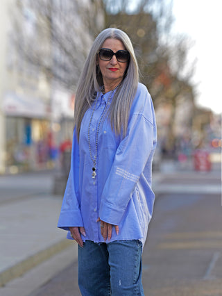 Nuomeno Concept Bluse Azzurro, getragen von einer Frau mit Sonnenbrille. Die Bluse hat eine Knopfleiste und eine gedrehte Brusttasche, hinten länger geschnitten mit asymmetrischer Rückenfalte.