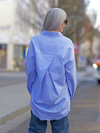 Frau trägt die Nuomeno Concept Bluse Azzurro mit Knopfleiste und gedrehter Brusttasche, hinten länger geschnitten, in einer Straßenumgebung. Ideal für stilvolle Alltagslooks.