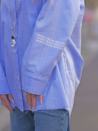Person trägt die Nuomeno Concept Bluse Azzurro mit Knopfleiste und gedrehter Brusttasche, hinten länger geschnitten, asymmetrische Falte am Rücken, aus 100% Baumwolle.